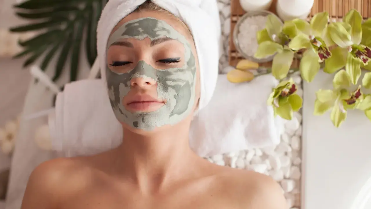 Femme détendue avec un masque fait maison, enveloppée dans une serviette, entourée de fleurs d'orchidée et de bougies.