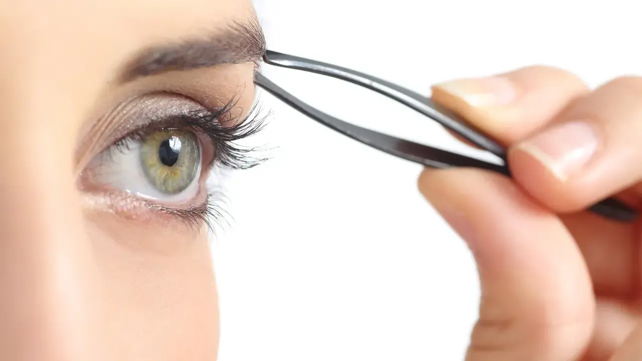 Une main utilise une pince à épiler pour parfaire l'épilation des sourcils d'un œil vert aux longs cils.