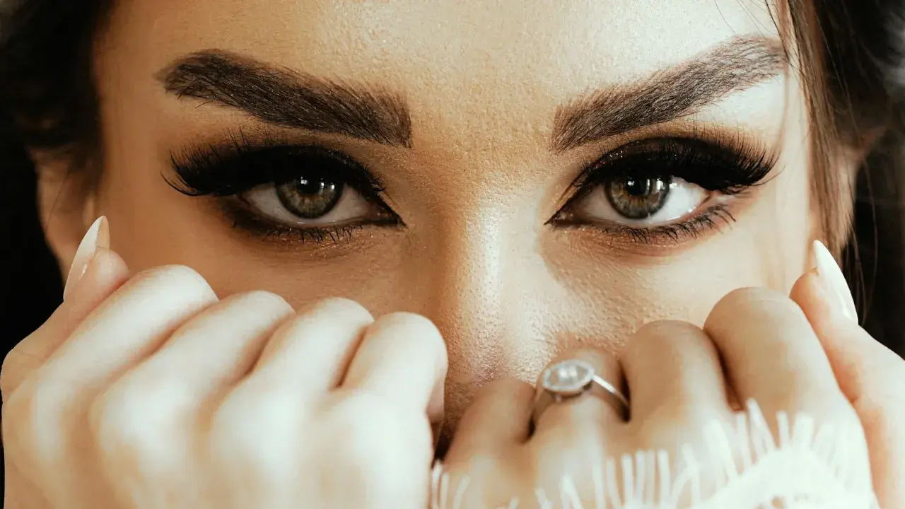 Yeux verts intenses, cils longs et maquillage sourcils parfait. Mains délicates ornées de bagues cachent une partie du visage.