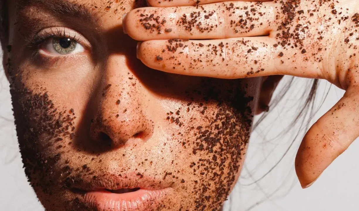 Une main couverte de grains de café recouvre un œil, le visage est enduit d'un gommage.
