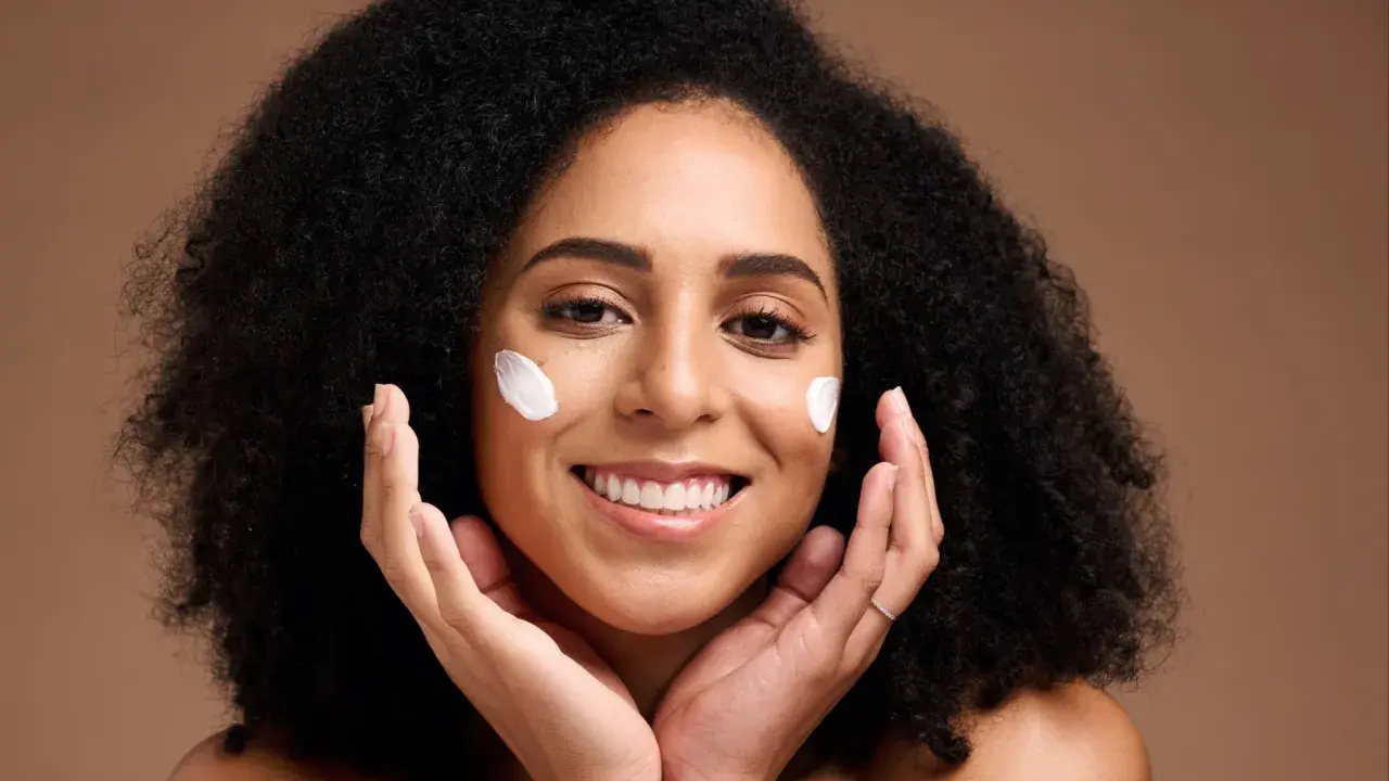 Une femme souriante applique du beurre de karité visage sur ses joues. Ses mains caressent doucement sa peau.