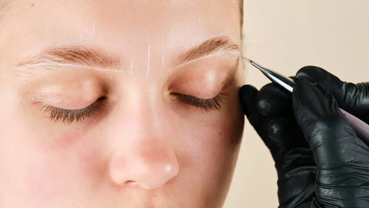 Une main gantée dessine des lignes blanches sur un sourcil pour un microblading poil à poil.