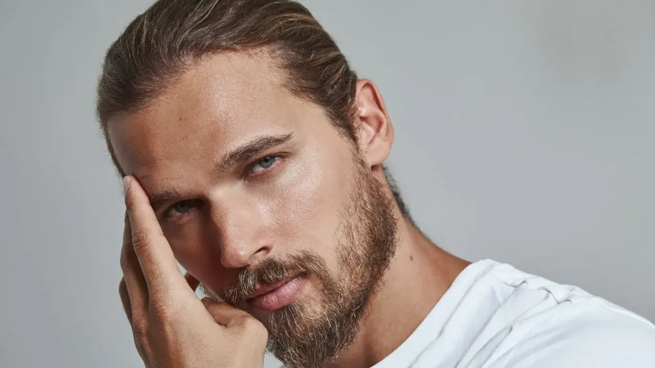 Un homme pensif, sa main effleurant son front, un sourcil plus haut que l'autre, regard intense.