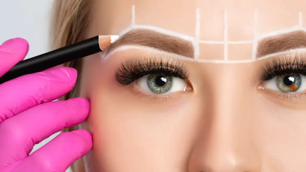 Main d'une esthéticienne en gant rose dessinant la forme sourcil visage rond avec un crayon blanc.