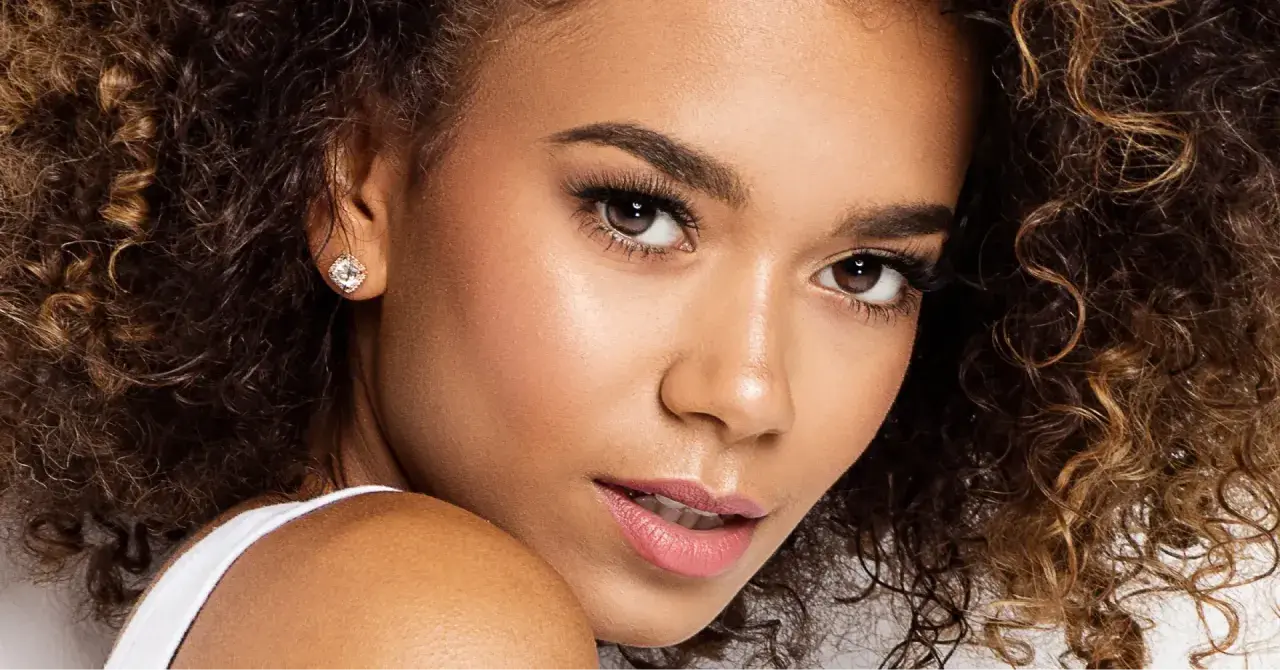 Un gros plan sur le visage d'une jeune femme aux cheveux bouclés, portant une boucle d'oreille étincelante. Ses **rehaucils avis** sont subtils, accentuant son regard.