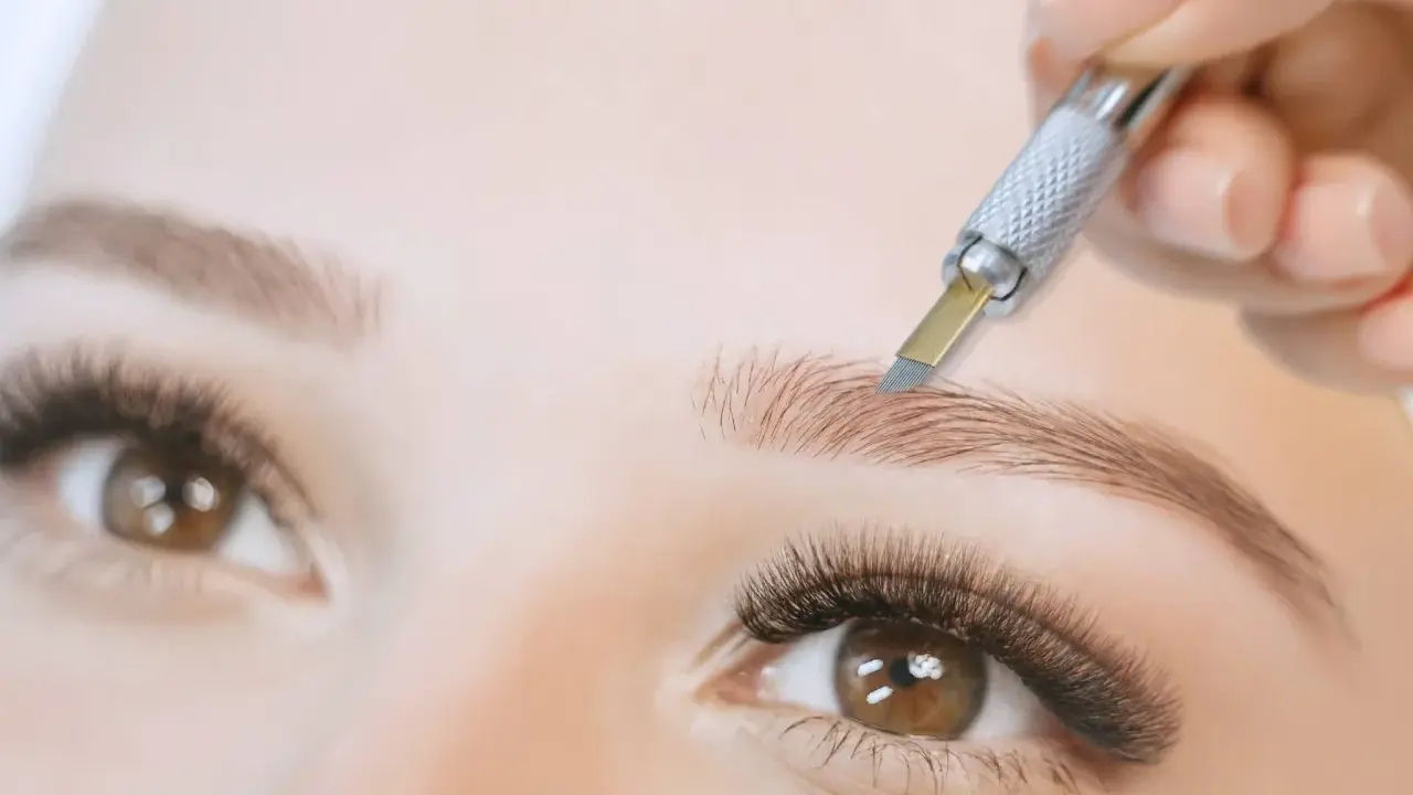 Main d'une esthéticienne réalisant un **tatouage sourcils poil par poil** avec un outil fin. Gros plan sur un œil et un sourcil.