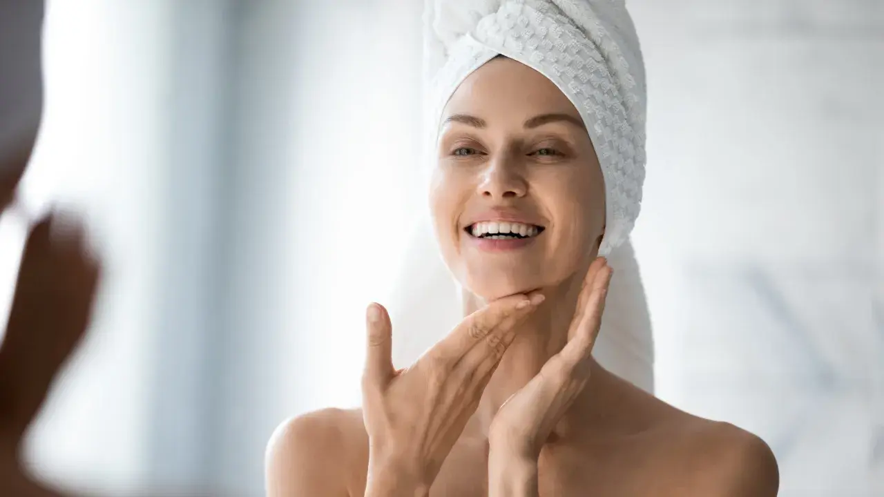 Une femme souriante, serviette sur la tête, applique délicatement de l'eau micellaire sur son visage pour une peau fraîche et nette.