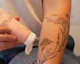 Une main tient un tube de crème réparatrice post-tatouage, prêt à être appliqué sur un bras orné d'un tatouage floral délicat.