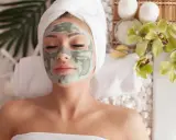 Femme détendue avec un masque fait maison, enveloppée dans une serviette, entourée de fleurs d'orchidée et de bougies.