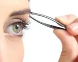 Une main utilise une pince à épiler pour parfaire l'épilation des sourcils d'un œil vert aux longs cils.