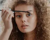 Une jeune femme aux cheveux bouclés applique un produit sur ses sourcils décolorés avec une brosse.