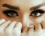 Yeux verts intenses, cils longs et maquillage sourcils parfait. Mains délicates ornées de bagues cachent une partie du visage.