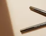 Deux crayons pour les yeux L'Atelier du Sourcil, un avant et un après, sur fond beige.
