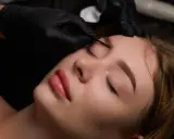 Une esthéticienne réalise un microblading sourcils sur une cliente allongée. Les mains gantées manipulent l'outil avec précision.