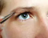 Une main utilise une pince à épiler pour affiner des sourcils, créant une ligne élégante au-dessus d'un œil bleu-vert.