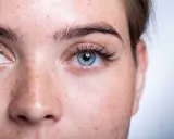 Gros plan sur un visage de femme aux yeux bleus et à la peau tachetée de rousseur. Ses cils courts et peu fournis encadrent un regard intense.