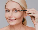 Une femme blonde applique du maquillage sur son œil, son sourcil blanc est visible.