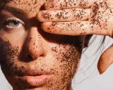 Une main couverte de grains de café recouvre un œil, le visage est enduit d'un gommage.