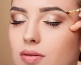 Une main applique un maquillage subtil pour parfaire la forme sourcil d'une femme aux yeux fermés.
