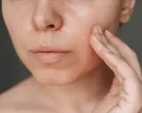 Une main touche délicatement la joue d'une femme, révélant une peau lumineuse, idéale pour une routine peau grasse.