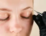 Une main gantée dessine des lignes blanches sur un sourcil pour un microblading poil à poil.