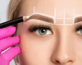 Main d'une esthéticienne en gant rose dessinant la forme sourcil visage rond avec un crayon blanc.