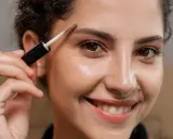 Une femme souriante utilise une brosse pour redessiner ses sourcils.