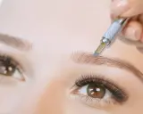 Main d'une esthéticienne réalisant un **tatouage sourcils poil par poil** avec un outil fin. Gros plan sur un œil et un sourcil.