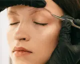 Une main gantée retire délicatement un poil incarné au sourcil d'une femme aux yeux fermés.