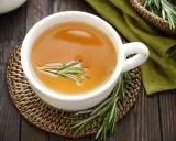Une tasse d'infusion romarin dorée, garnie d'une branche de romarin frais, sur un dessous de plat en osier.