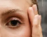 Un œil attentif, cils longs, et le début du repousse sourcils après chimio. Une main effleure le visage.