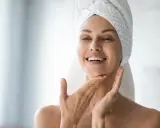 Une femme souriante, serviette sur la tête, applique délicatement de l'eau micellaire sur son visage pour une peau fraîche et nette.