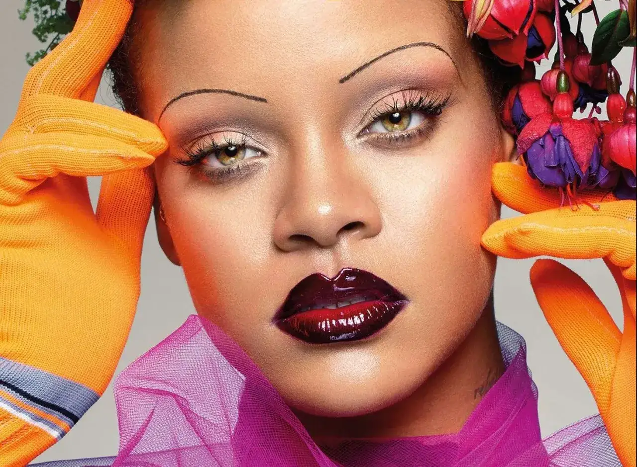 Un sourcil fin, style année 2000, encadre un regard intense. Lèvres sombres et brillantes, gants orange vifs, fleurs colorées.