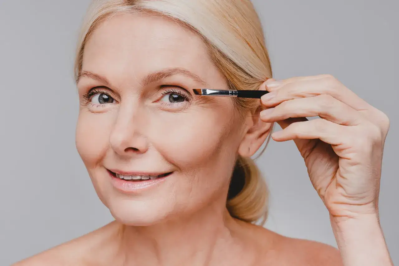 Une femme blonde applique du maquillage sur son œil avec un pinceau, accentuant son sourcil blanc.