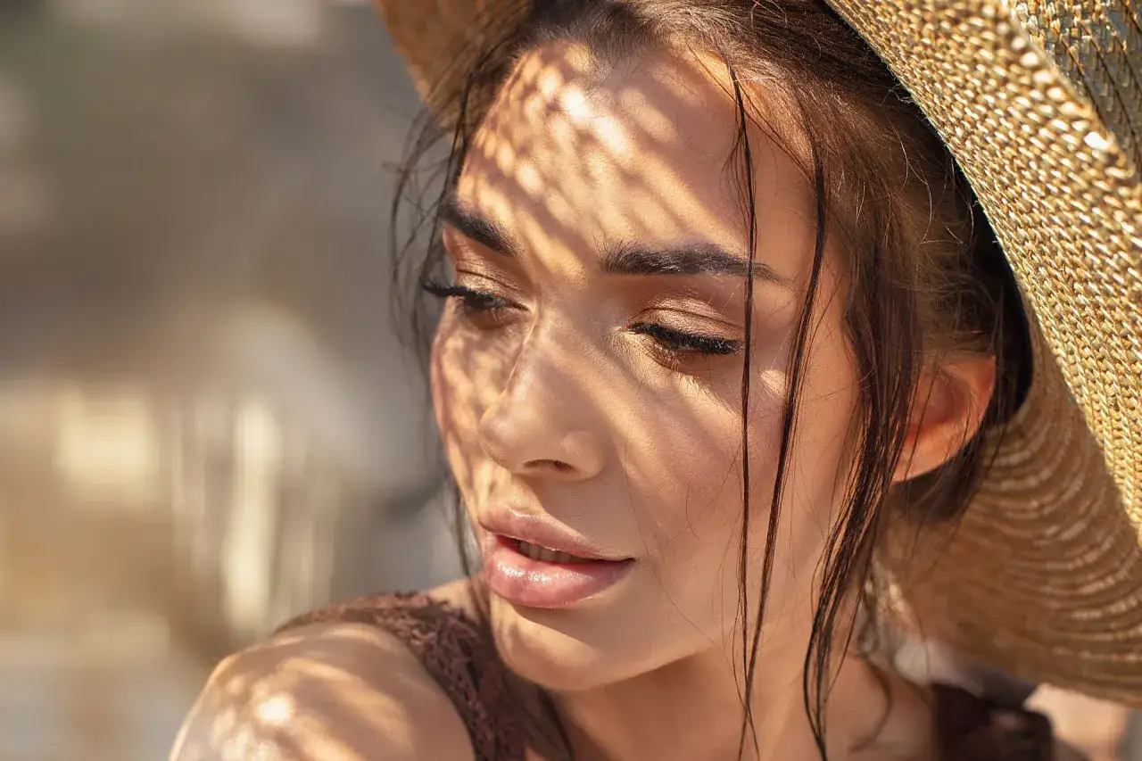 Visage d'une femme sous un chapeau de paille, ombres du soleil sur sa peau. Ses sourcils, sublimés par le microblading, ressortent parfaitement.