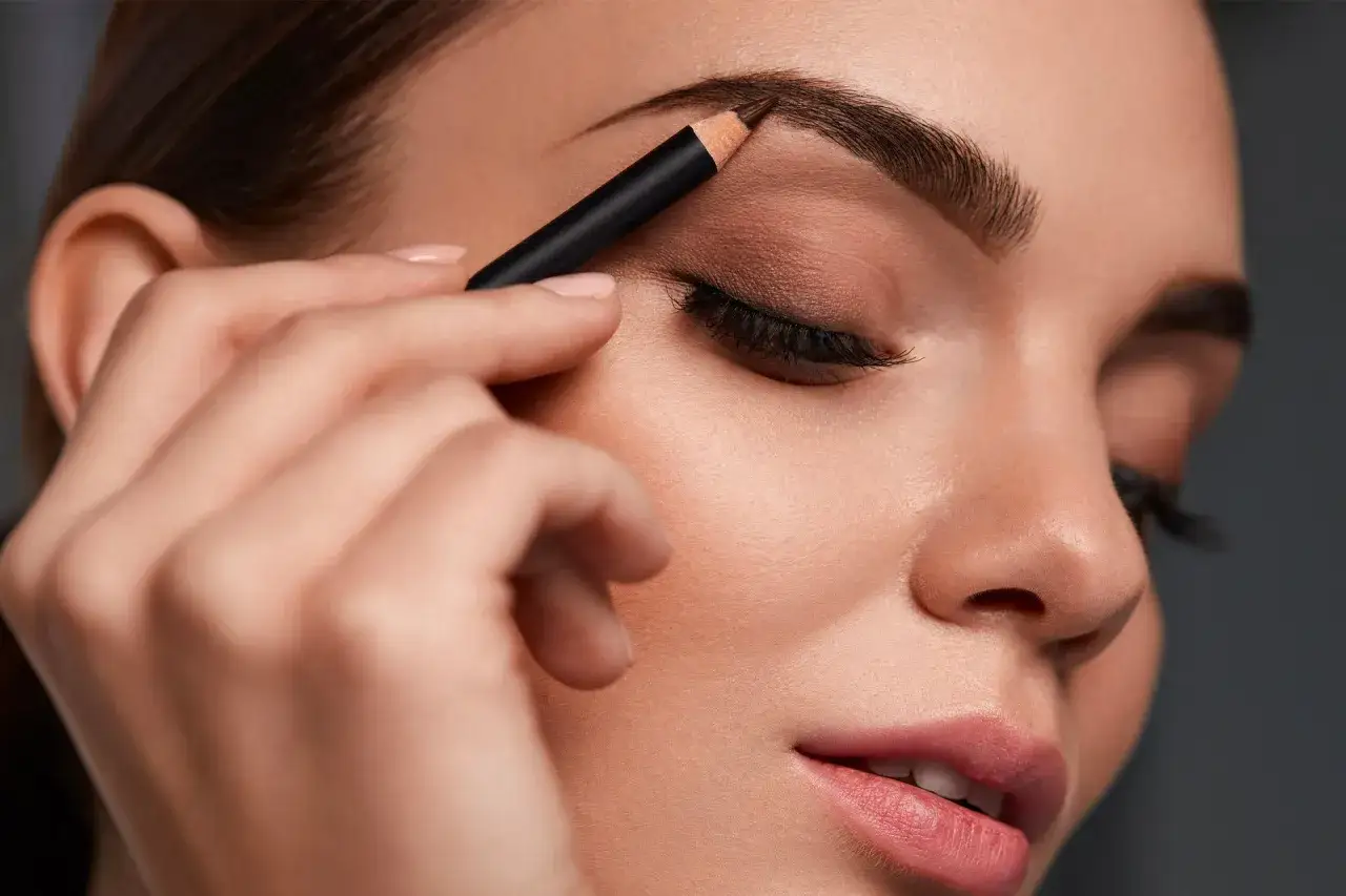 Une main applique un crayon pour redessiner des sourcils fins, accentuant la beauté du regard.