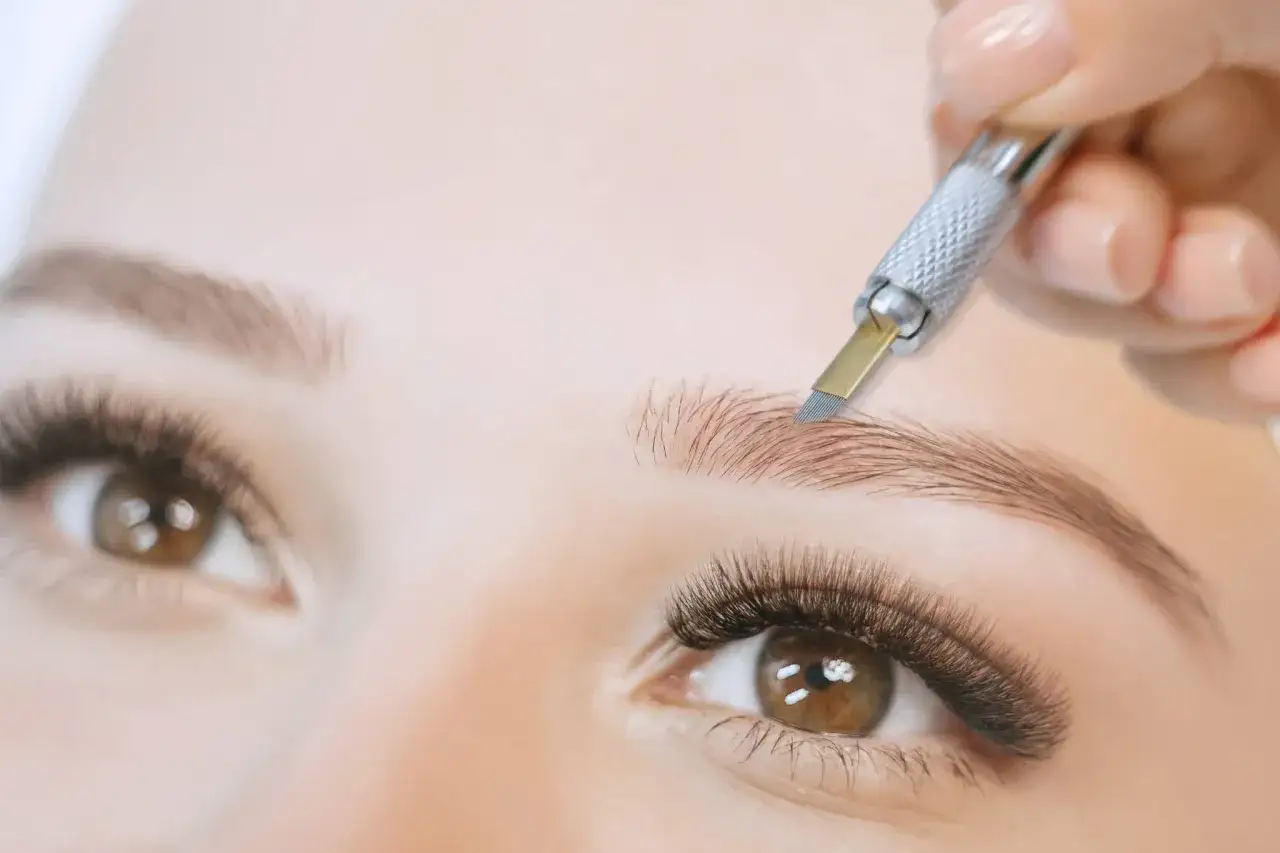 Main d'une esthéticienne réalisant un tatouage sourcils poil par poil avec un outil fin.