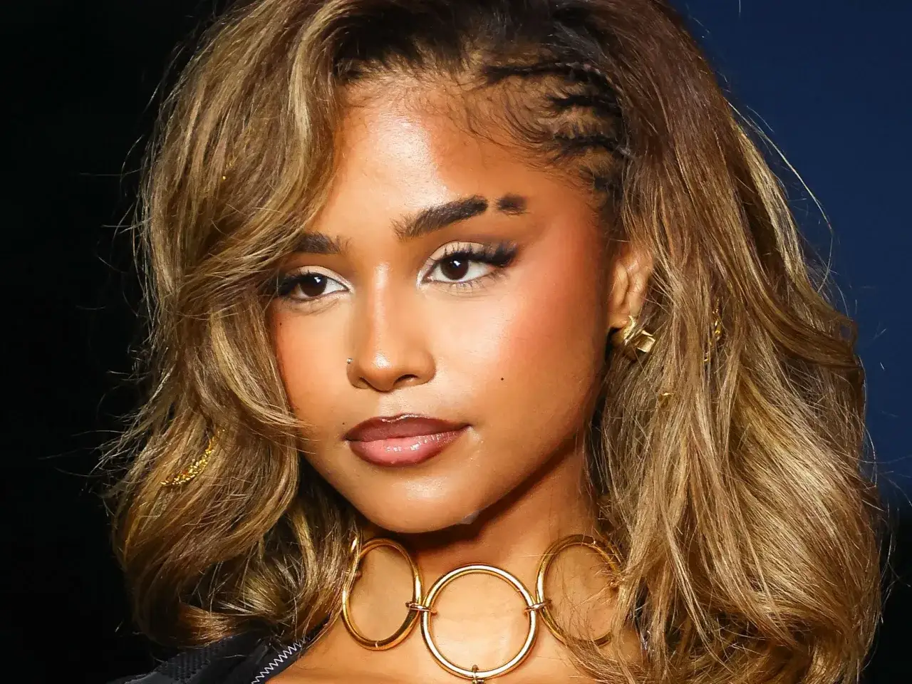Un sourcil coupé femme, orné de tresses sur le côté, arbore un maquillage audacieux et un collier doré.